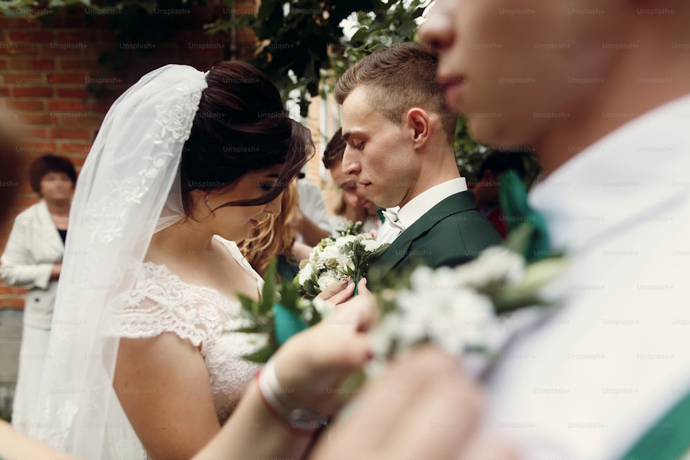 Schöne Braut im Vintage-Hochzeitskleid, die vor der Zeremonie Boutonniere auf den hübschen Bräutigam und seine stilvollen Trauzeugen im Freien legt, Hände und Blumen Nahaufnahme