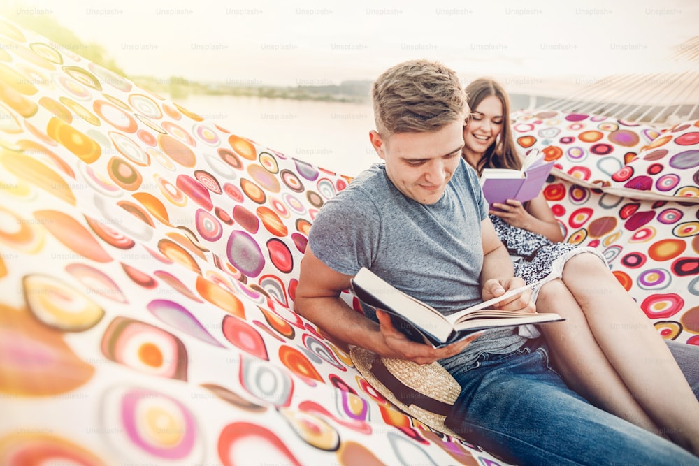 행복한 힙스터 커플은 책을 읽고 해변에서 일몰 햇살을 받으며 해먹에서 휴식을 취하고, 여름 휴가를 보내고 있습니다. 책을 들고 여름 저녁에 쉬는 세련된 힙스터. 텍스트를 위한 공간