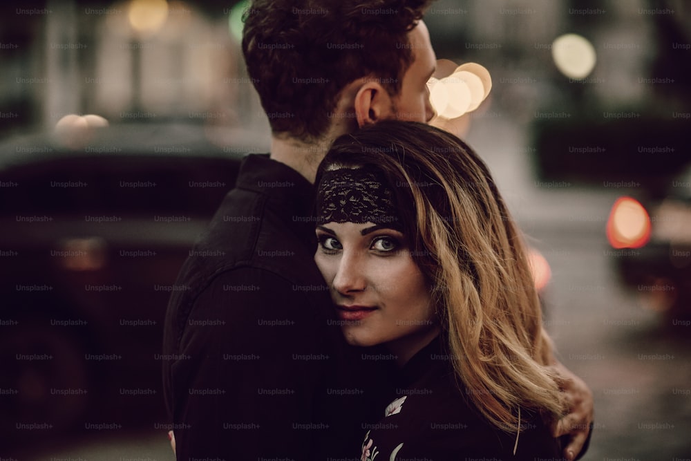 スタイリッシュなジプシーのカップルが、動く車のライトで夜の街の通りで抱きしめています。女性と男性が抱き合う、ロマンチックなフランスの雰囲気の瞬間。情熱的な愛の気分。