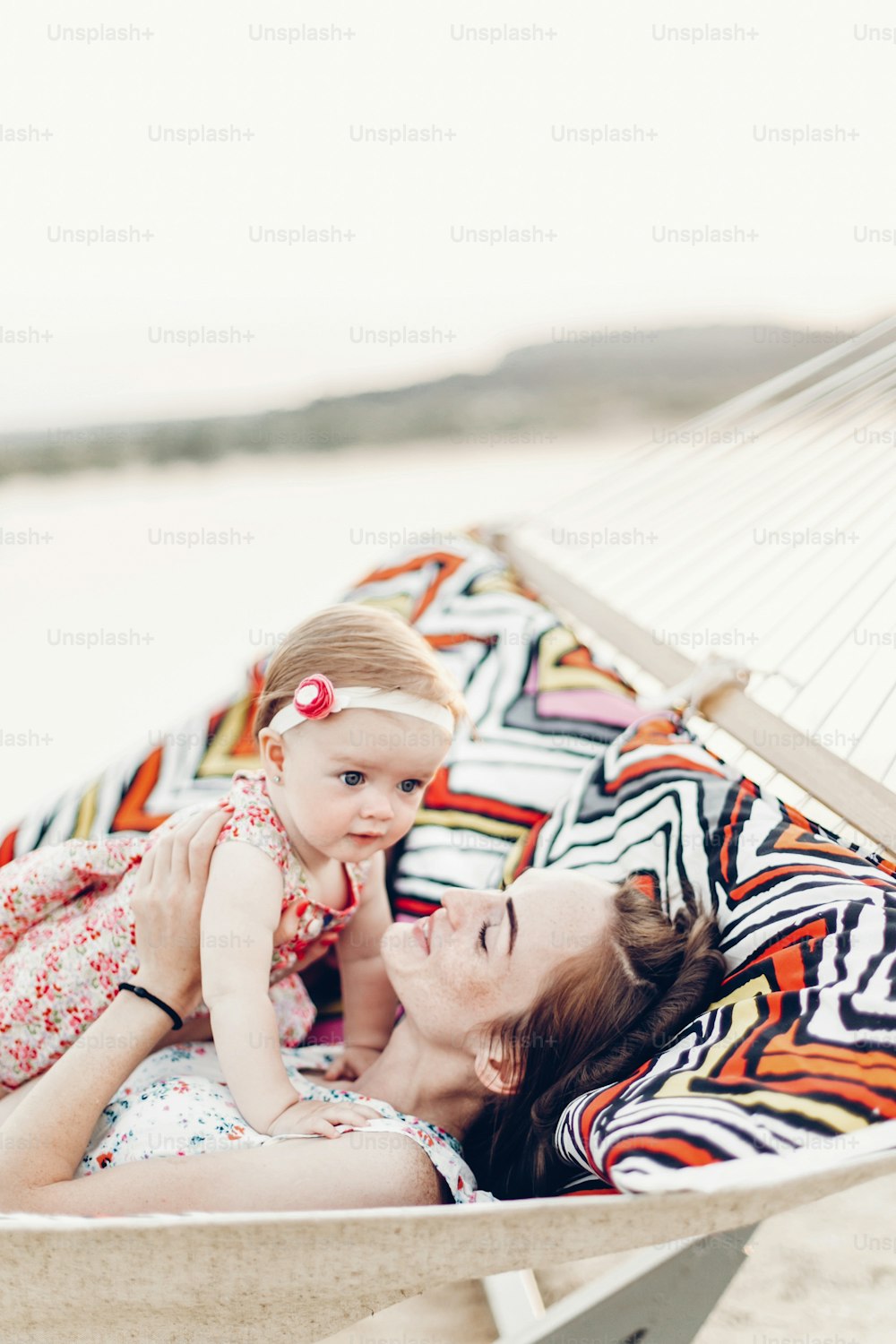 Happy young mother holding baby daughter in hands while relaxing in hammock with husband, hipster family concept, cute girl with her mommy, close-up