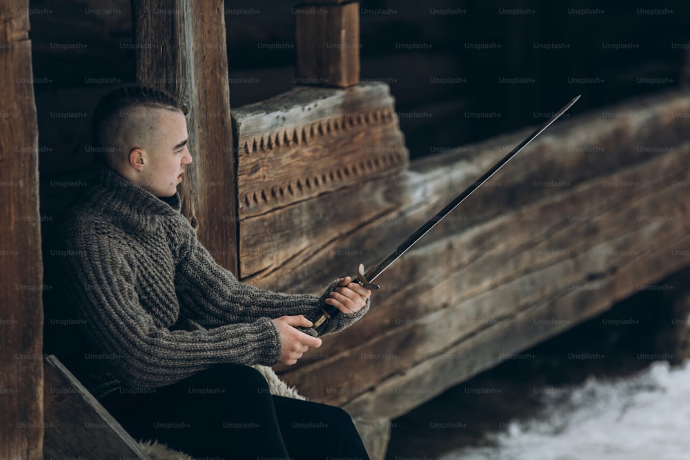 Brave warrior holding sword near historical wood castle building in winter woods in Scandinavia, handsome viking with mohawk posing with weapon before battle, fantasy cosplay concept