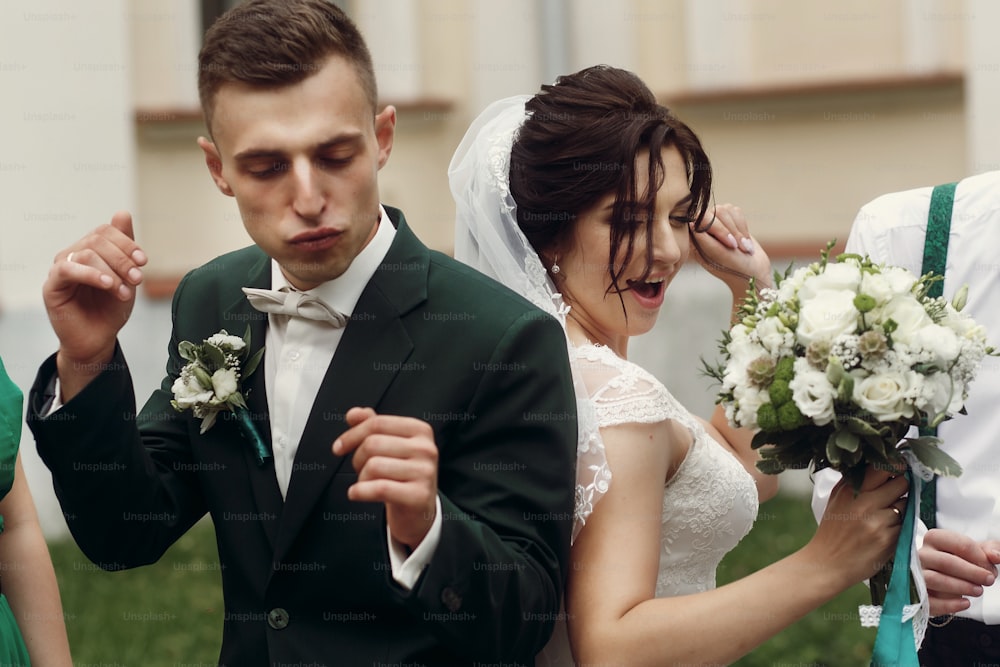 Glückliches frisch verheiratetes Paar, lächelnde brünette Braut mit Blumenstrauß und hübschem stilvollem Bräutigam, der tanzt und Spaß im Freien im Park mit ihren Freunden hat