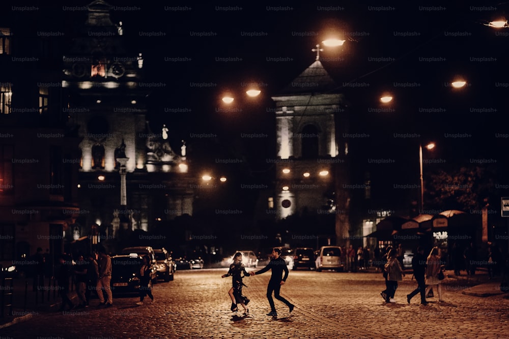 lovers running through empty road in the evening city street. stylish couple in love having fun and holding hands in city lights. modern woman and man romantic french atmospheric moment