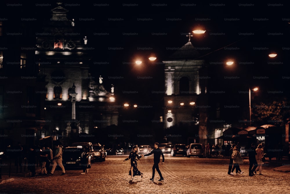 Coppia felice su una passeggiata in strada europea durante la loro luna di miele, bella donna in abito nero che si tiene per mano con un bell'uomo in abito nero all'aperto nella notte, concetto di avventura