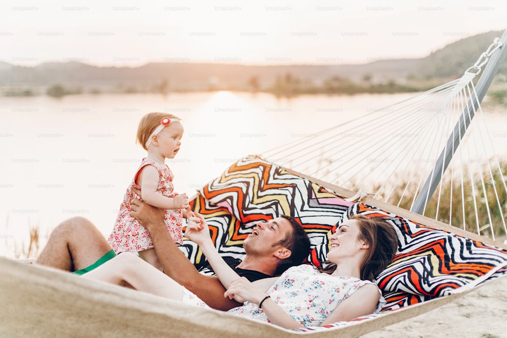 Young family with baby on the beach, smiling father and mother holding cute little girl while lying in a hammock, hipster family on vacation near lake at sunset