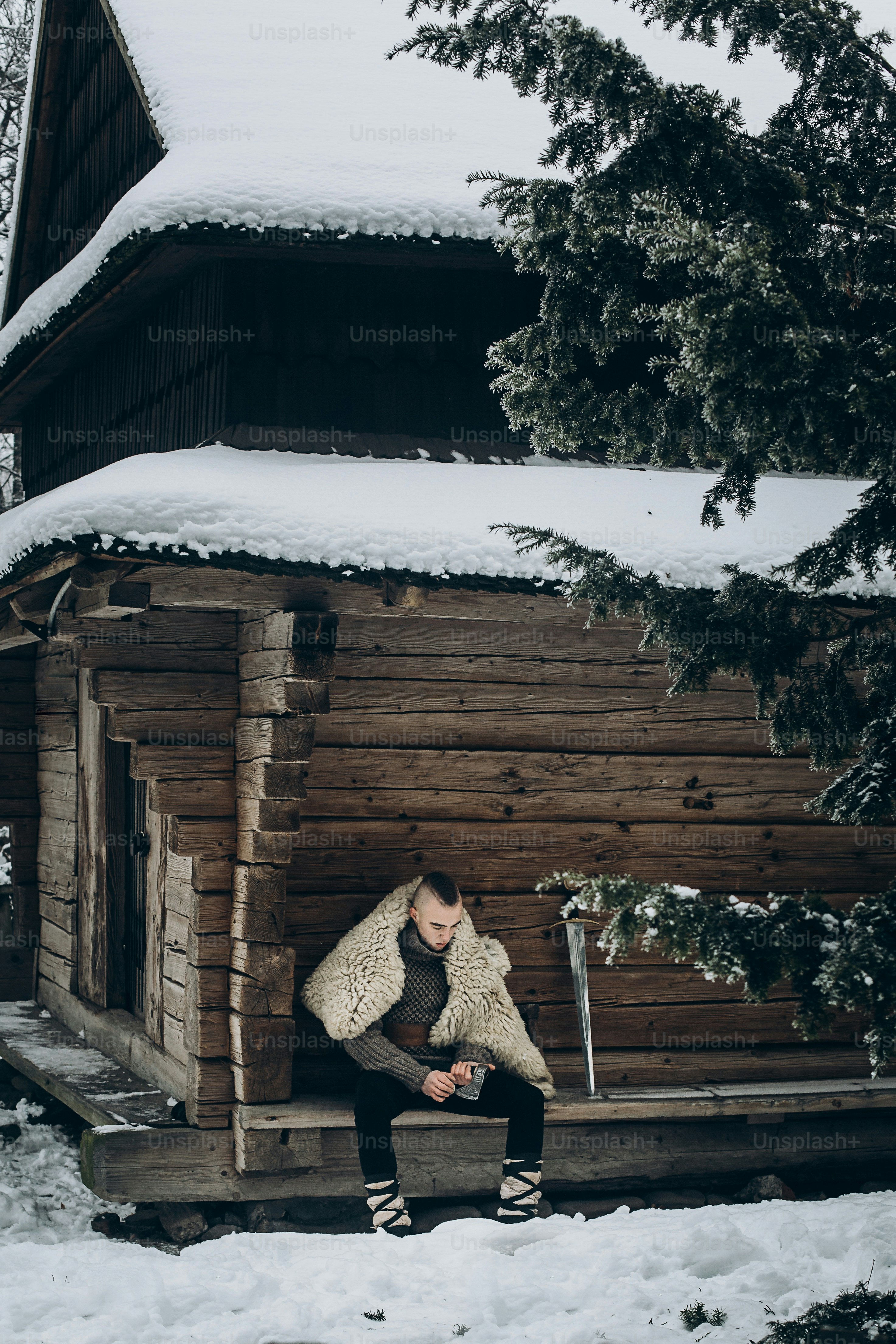 Brave viking warrior sharpening axe before battle in the north, scandinavian man with mohawk sitting near historical wooden building near sword, viking cosplay concept
