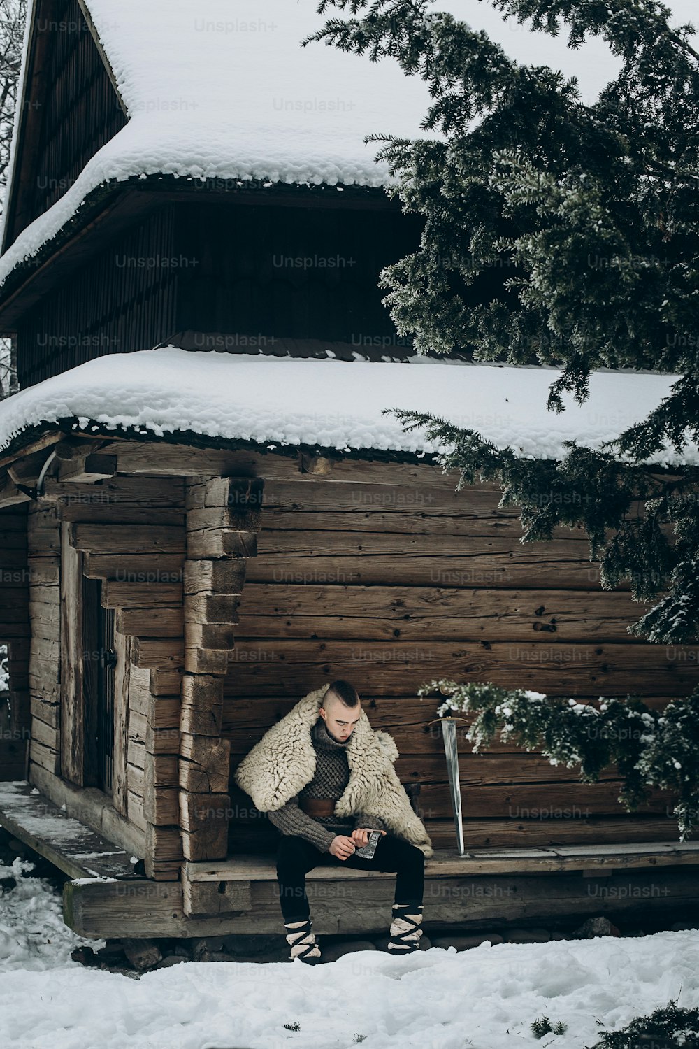 Tapferer Wikingerkrieger schärft Axt vor der Schlacht im Norden, Skandinavier mit Mohawk sitzt in der Nähe eines historischen Holzgebäudes in der Nähe von Schwert, Wikinger-Cosplay-Konzept