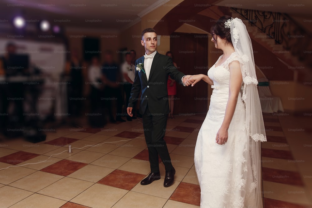 Pareja romántica de recién casados bailando, novio guapo y novia hermosa feliz bailan primero mientras se toman de la mano en la recepción de la boda del restaurante, hermosa iluminación