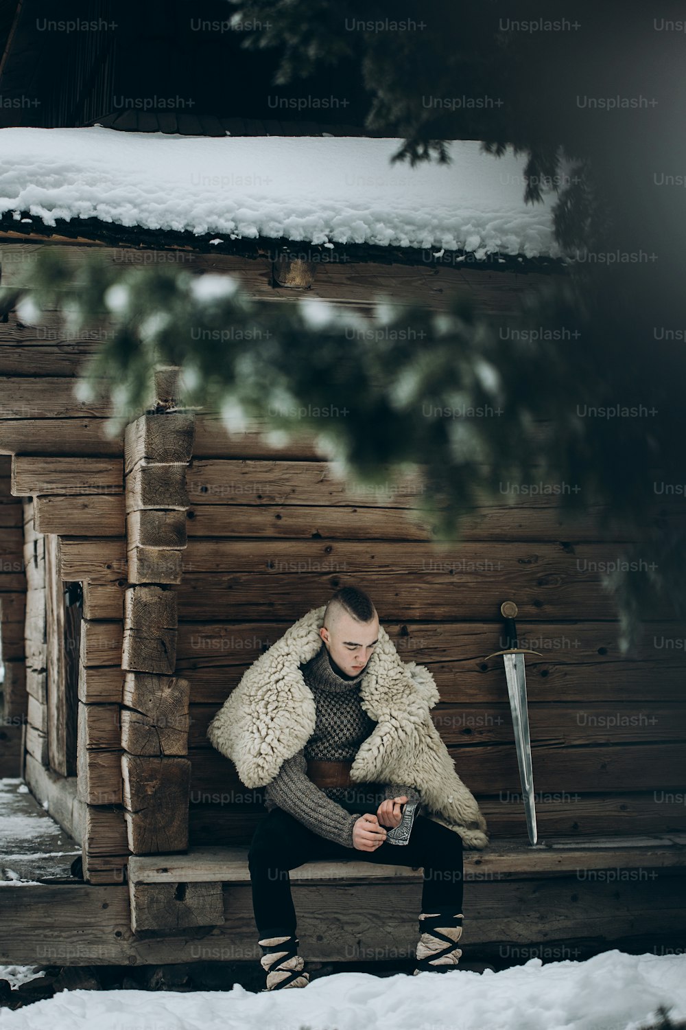 Tapferer Wikingerkrieger schärft Axt vor der Schlacht im Norden, Skandinavier mit Mohawk sitzt in der Nähe eines historischen Holzgebäudes in der Nähe von Schwert, Wikinger-Cosplay-Konzept
