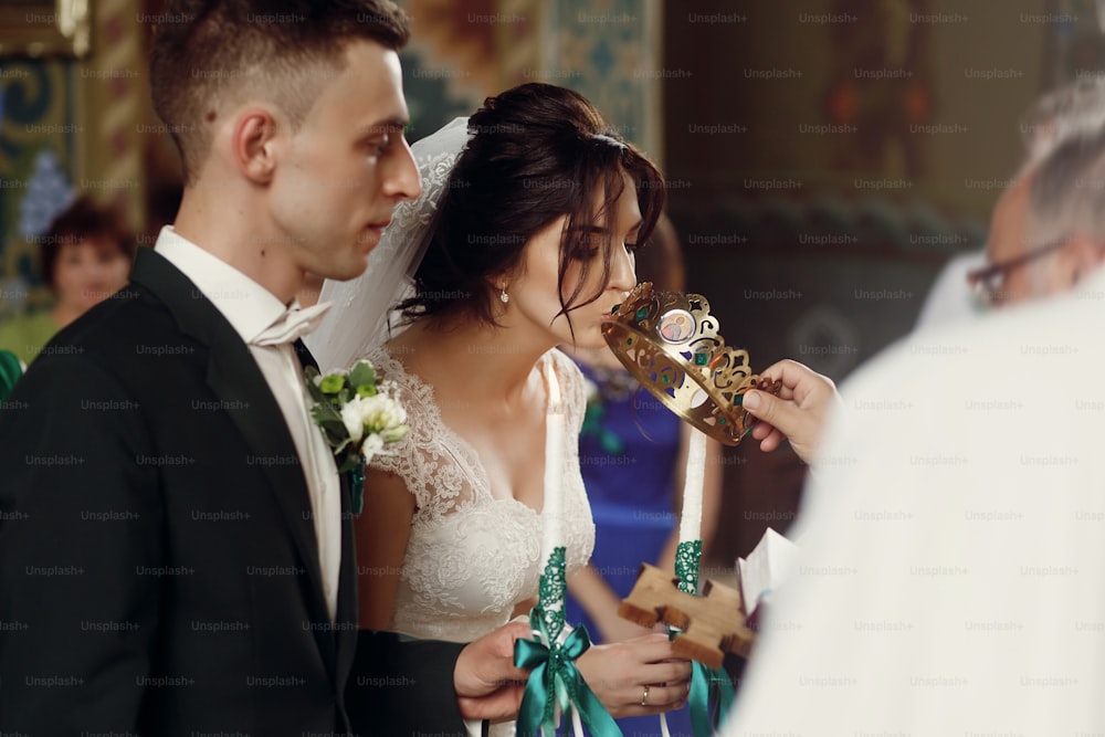 Happy spiritual couple, stylish groom and beautiful brunette bride in white dress holding candles at wedding church ceremony during golden crown coronation