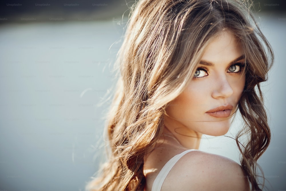 beautiful boho girl portrait with amazing eyes and lips standing near lake. attractive young woman in white bohemian dress with windy hair posing on sand beach near water