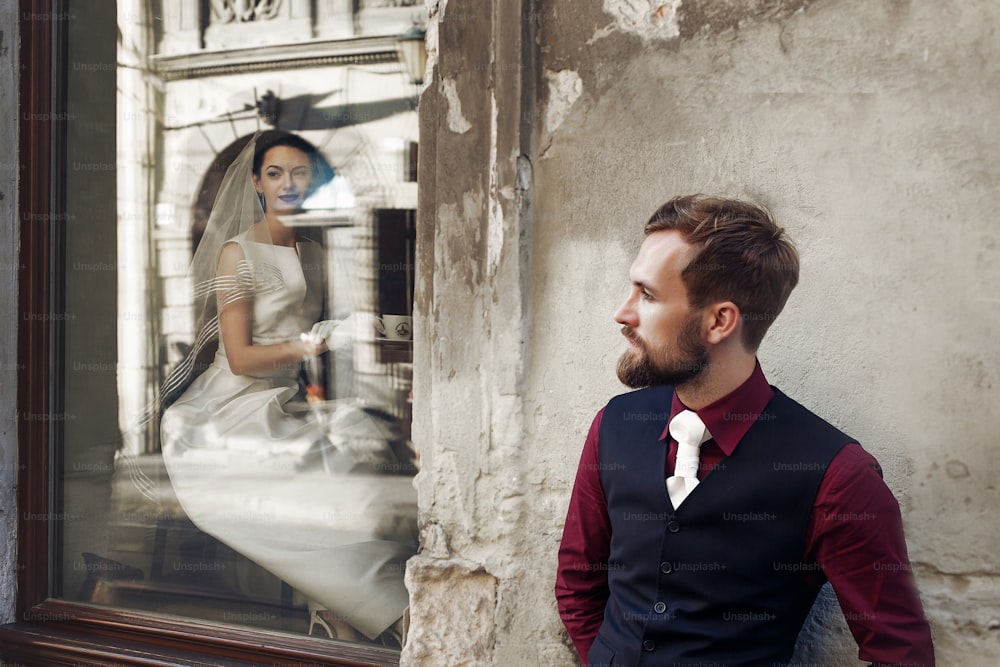 elegant gorgeous bride  drinking coffee in window and stylish groom posing at building in street outdoors cafe. unusual luxury wedding couple in retro style. romantic moment
