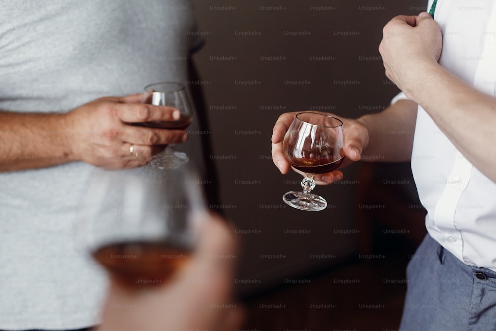 Handsome groom and his groomsman friends in stylish suits drink whiskey in hotel room, morning before the wedding preparation, hands closeup