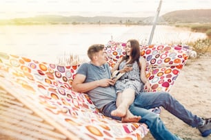 happy couple looking at phones and smiling, relaxing in hammock in sunset light on the beach. stylish hipsters having fun, and resting on summer vacation. social media concept, space for text