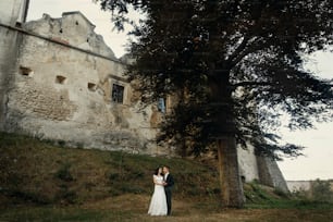 スタイリッシュで豪華な新郎新婦が夕暮れ時に古城の近くで一緒にポーズをとっています。屋外の美しい結婚式のカップルの幸せな瞬間