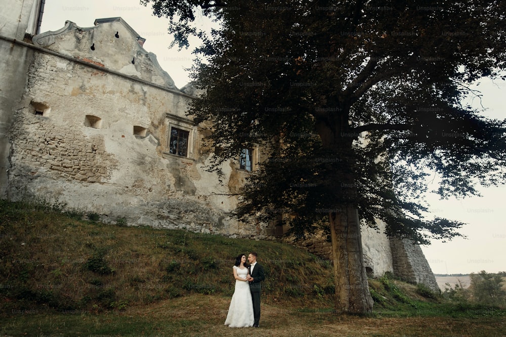 stylish luxury bride and groom posing together near old castle at sunset. happy moment of beautiful wedding couple outdoors