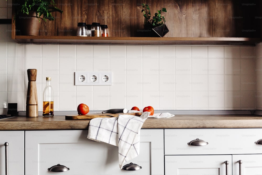 Tabla de madera con cuchillo, tomates, aceite de oliva en encimera de cocina moderna y estante con especias y plantas. cocinar alimentos. Elegante diseño interior de cocina en estilo escandinavo, electrodomésticos de acero