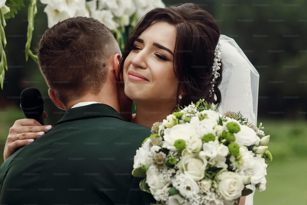 Gorgeous emotional bride in stylish white wedding dress with bouquet hugging handsome groom during outdoor wedding ceremony near aisle , her eyes closed, face closeup