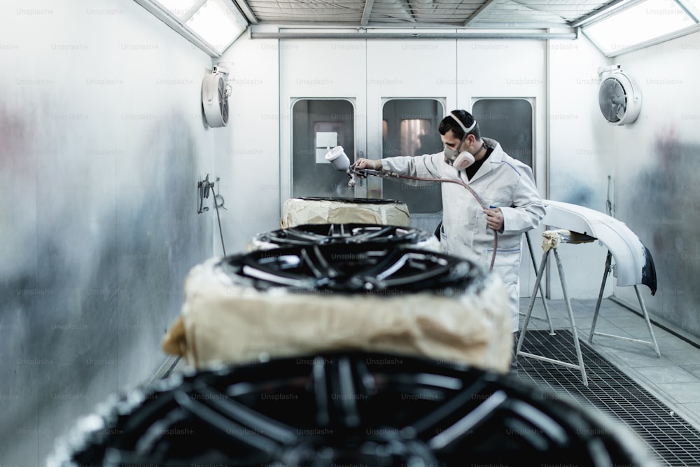 Man with protective clothes and mask painting car parts using spray compressor. Selective focus.