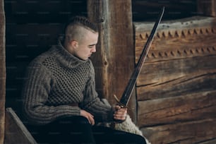 Valiente guerrero sosteniendo la espada cerca del edificio histórico del castillo de madera en los bosques de invierno en Escandinavia, vikingo guapo con mohawk posando con arma antes de la batalla, concepto de cosplay de fantasía