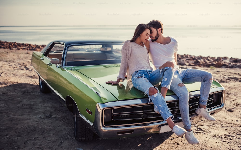 Romantic couple is sitting on green retro car on the beach. Handsome bearded man and attractive young woman with vintage classic car. Love story.