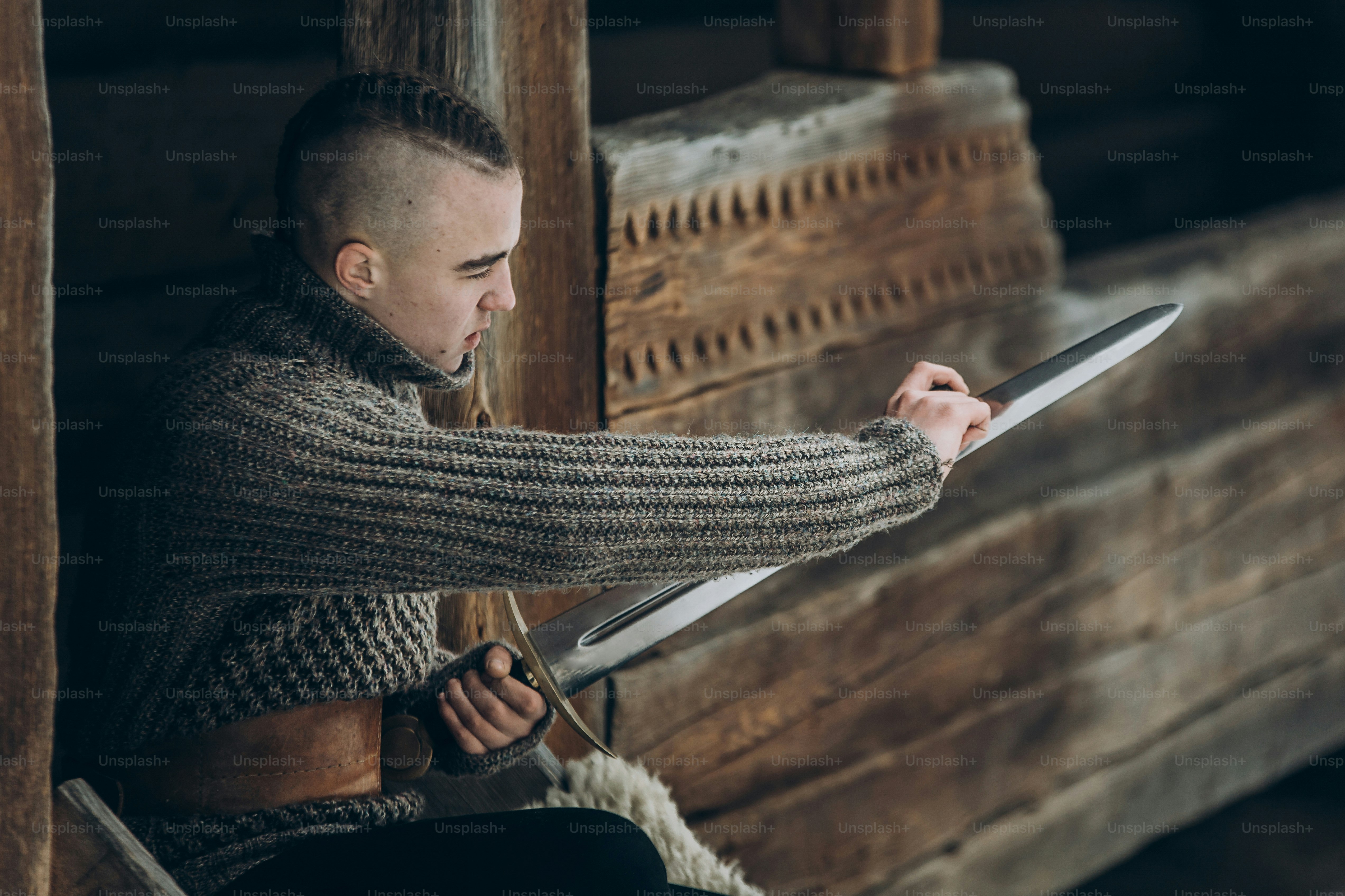 Strong viking warrior sharpening his sword while sitting near ancient wooden castle, scandinavian knight with weapon in viking costume, historical heritage concept