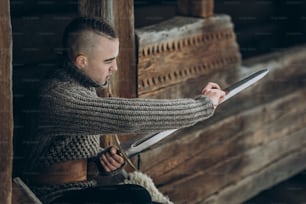 Strong viking warrior sharpening his sword while sitting near ancient wooden castle, scandinavian knight with weapon in viking costume, historical heritage concept