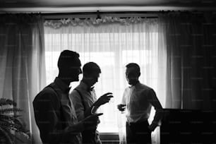 Handsome groom and his groomsman friends in stylish suits drink whiskey in hotel room, morning before the wedding preparation, emotional group of friends celebrating