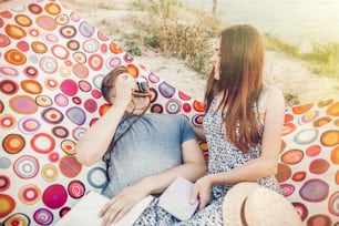 casal hipster feliz com livros e câmera fotográfica relaxando na rede à luz do pôr do sol na praia, férias de verão. homem elegante tirando foto de sua bela mulher. espaço para texto