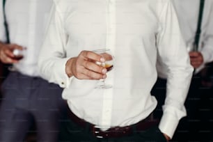 Stilvoller Bräutigam mit Trauzeugen, die morgens vor der Hochzeitsvorbereitung Whisky im Glas halten. Luxusgruppe von Freunden. Selbstbewusster Geschäftsmann
