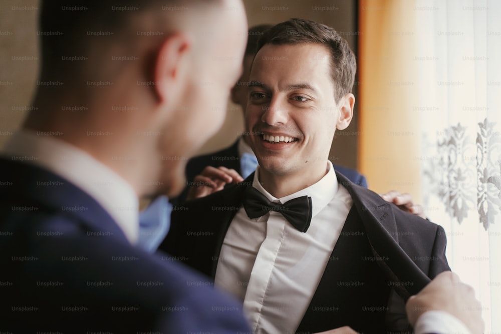 Groom getting ready in the morning with groomsmen in the room. man hands putting on jacket and bow tie, standing at window light in hotel. preparation for wedding day