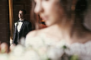 stylish groom looking at gorgeous bride in amazing dress with bouquet posing at window in luxury room in hotel. rich wedding couple. romantic moment of newlyweds in classic indoors