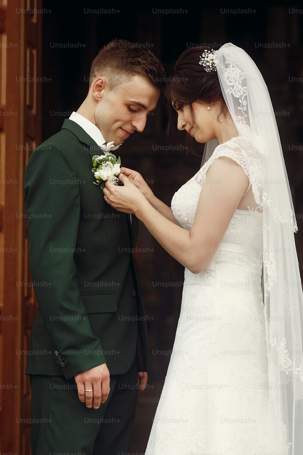 Happy couple of newlyweds, brunette bride in white wedding dress putting on boutonniere on handsome smiling groom, husband and wife at church doorway after ceremony