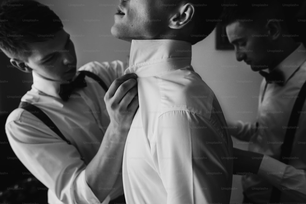 stylish handsome groom posing while groomsman putting on shirt and tying bow,  helping him to get ready in the morning. wedding preparation. black and white photo
