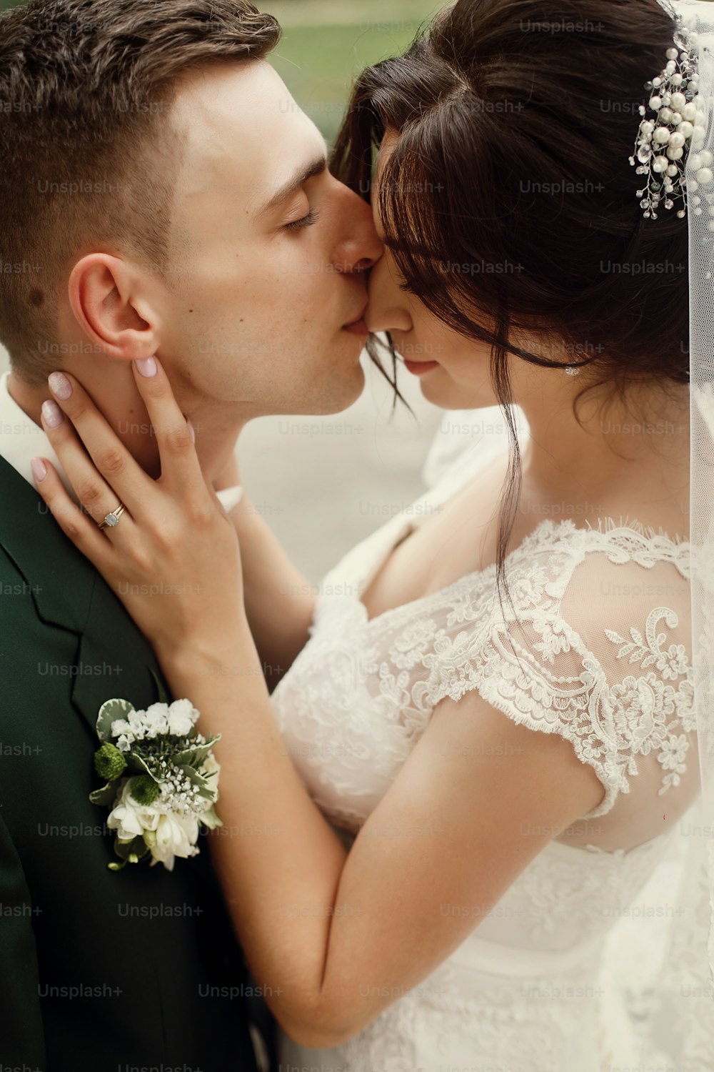 Gorgeous brunette bride in lace white wedding dress kissing handsome happy groom in stylish suit, newlyweds walking in park portrait, face closeup
