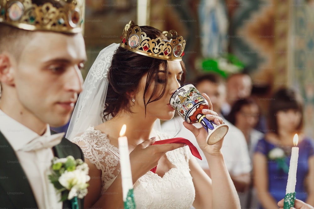 Young beautiful couple taking communion during wedding ceremony in christian church, beautiful bride in white dress and handsome groom in a crown holding candles, face closeup