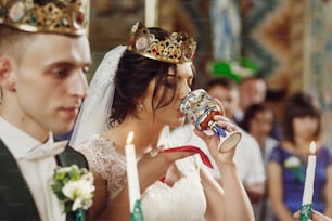 Young beautiful couple taking communion during wedding ceremony in christian church, beautiful bride in white dress and handsome groom in a crown holding candles, face closeup