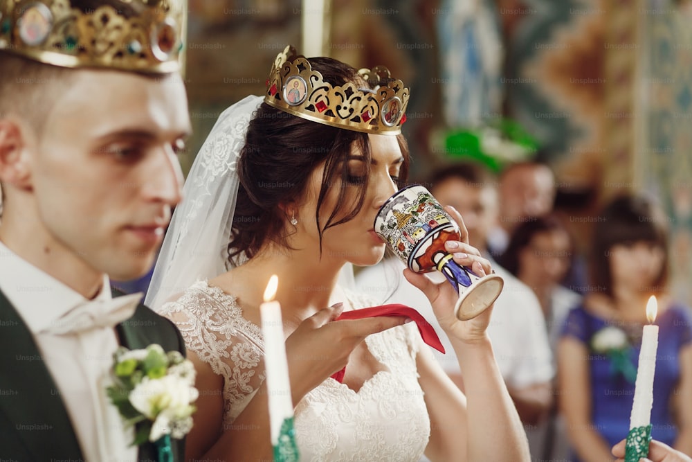 Jeune beau couple prenant la communion lors de la cérémonie de mariage dans l’église chrétienne, belle mariée en robe blanche et beau marié dans une couronne tenant des bougies, visage gros plan