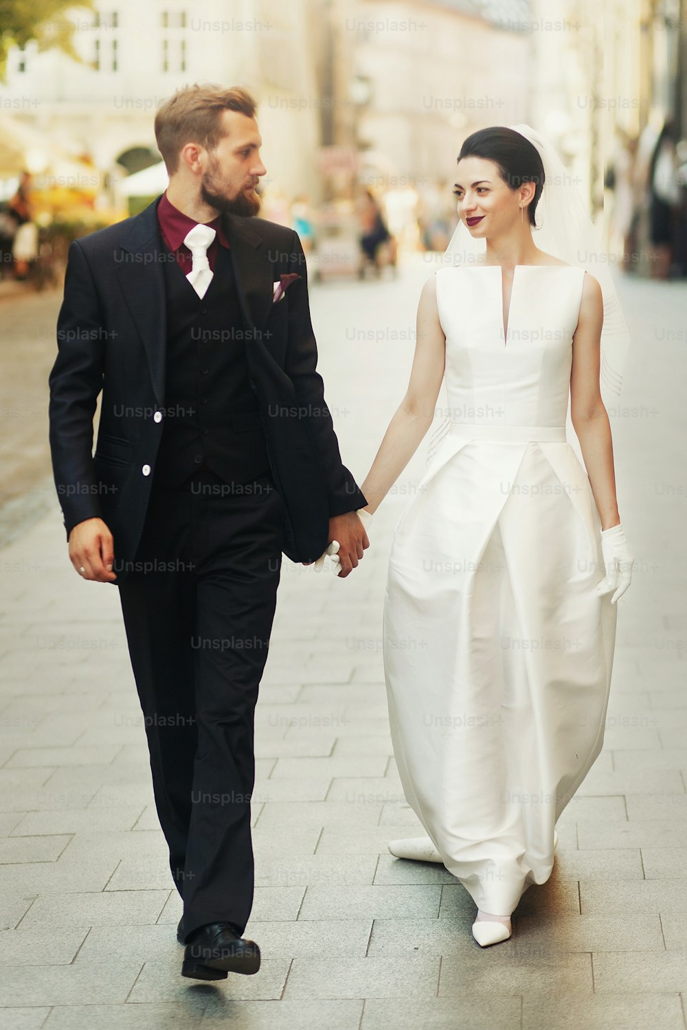 手をつないで街の通りを歩く豪華な結婚式のカップル。エレガントでゴージャスな花嫁とスタイリッシュな新郎の笑顔。レトロなスタイル。ロマンチックなひととき
