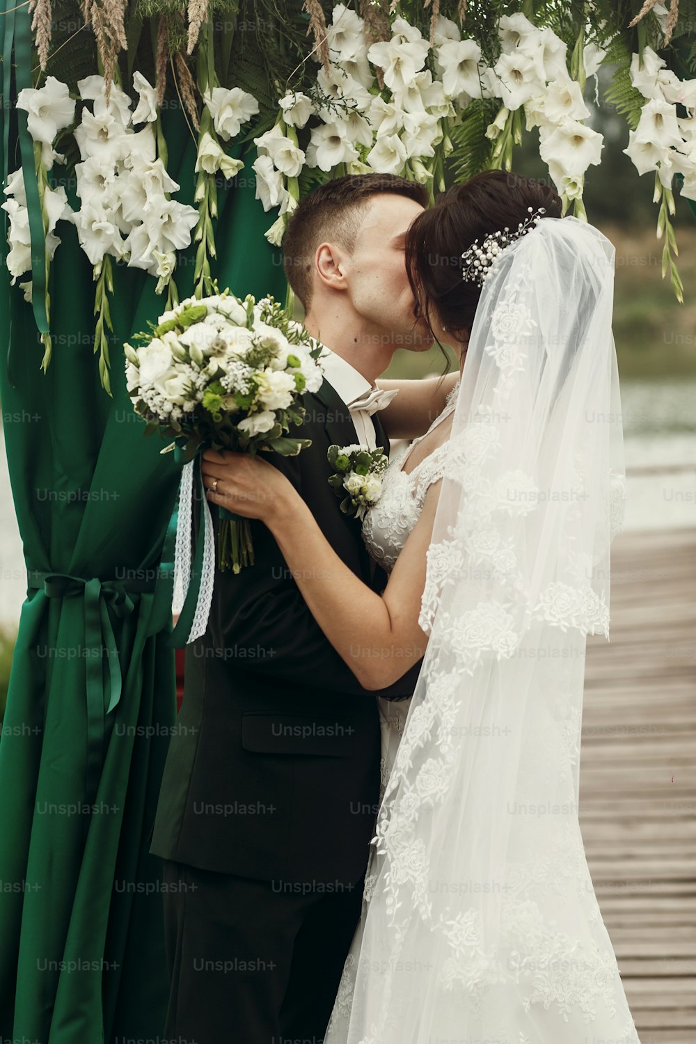 Happy newlywed couple hugging under white flowers aisle arch, handsome groom kissing beautiful brunette bride at wedding ceremony, family portrait