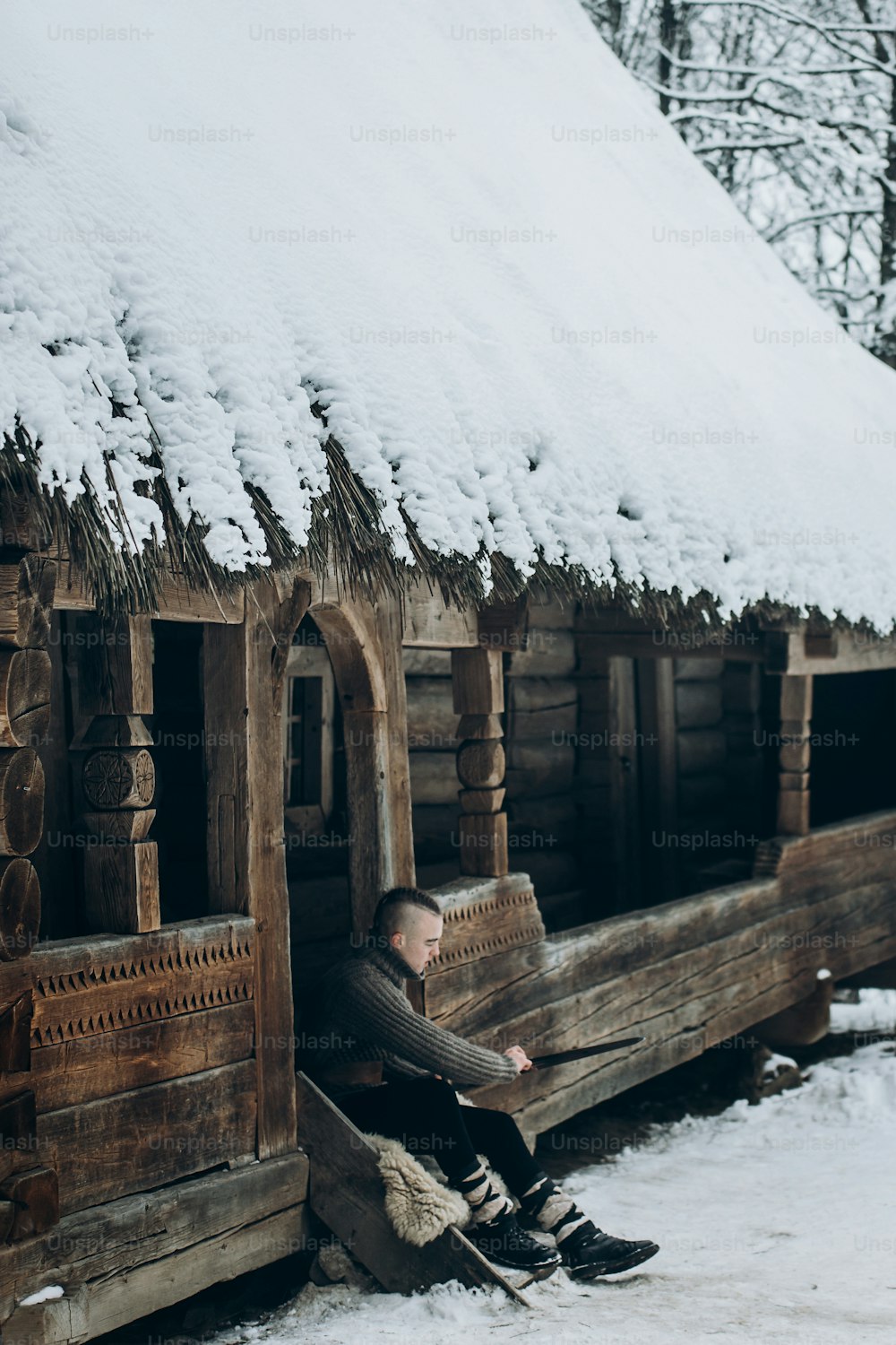 viking warrior sharpening his sword while sitting near ancient wooden castle, scandinavian knight with weapon in viking costume, historical heritage concept