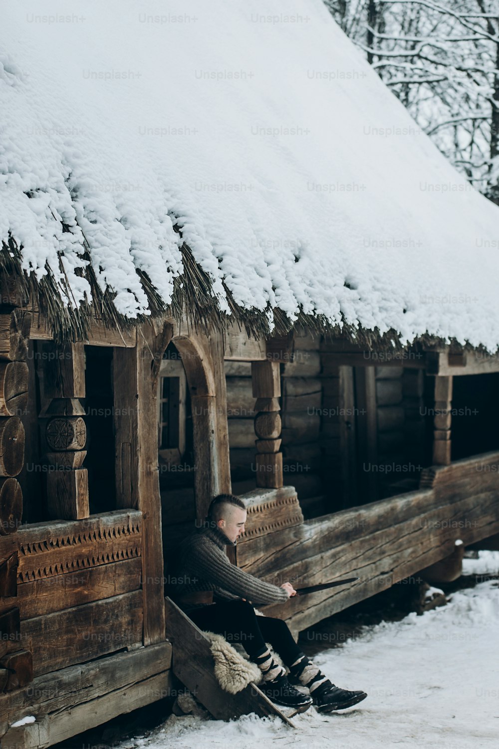 Wikingerkrieger schärft sein Schwert, während er in der Nähe einer alten Holzburg sitzt, skandinavischer Ritter mit Waffe im Wikingerkostüm, historisches Kulturerbekonzept