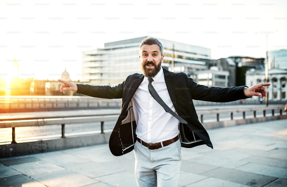 Alegre hombre de negocios hipster vestido con camisa blanca y chaqueta caminando por el puente de la ciudad, con los brazos extendidos. Concepto de celebración del éxito.