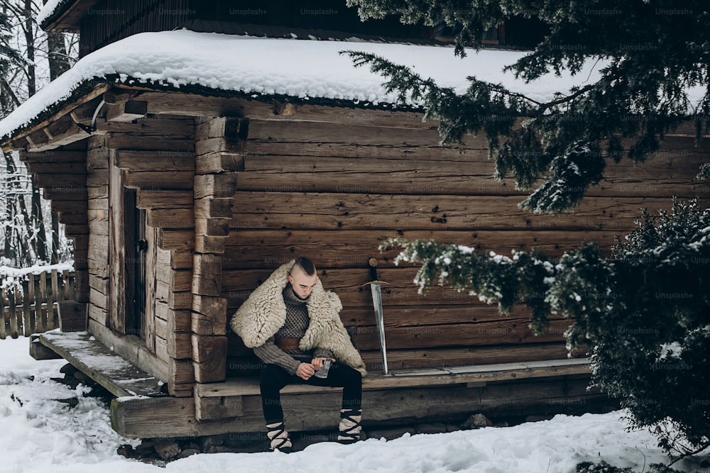 Brave viking warrior sharpening axe before battle in the north, scandinavian man with mohawk sitting near historical wooden building near sword, viking cosplay concept