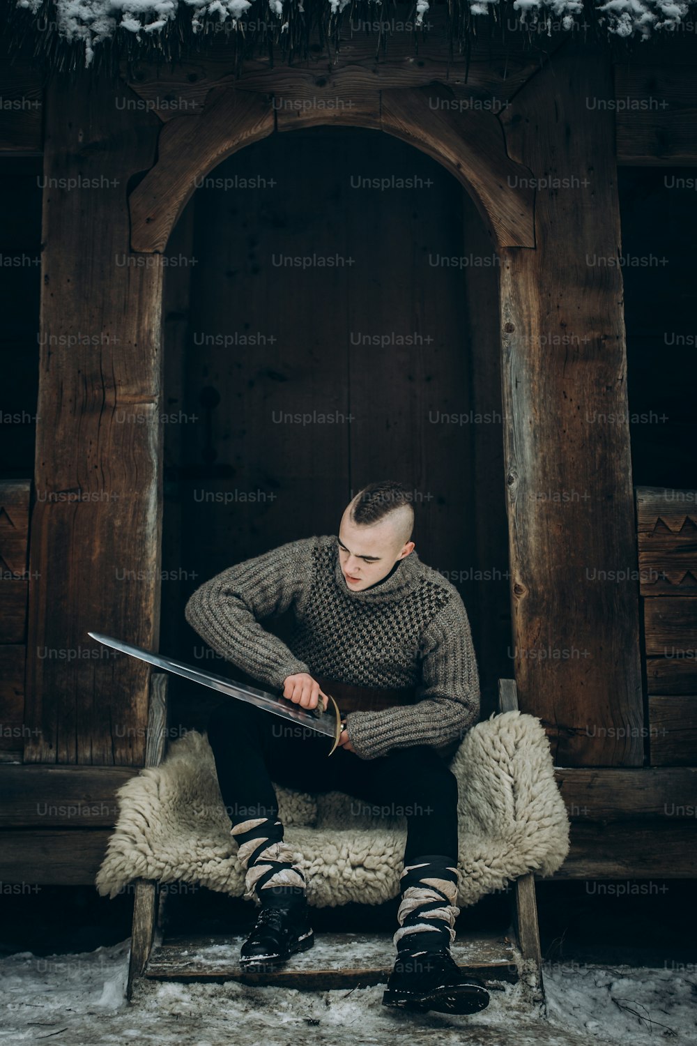 剣を研ぐ強いバイキングの戦士の肖像画、スカンジナビアのバイキングのコスプレ、冬の森のオオカミの毛皮に座っている鋼の剣を持つ中世の戦士、戦闘準備のコンセプト