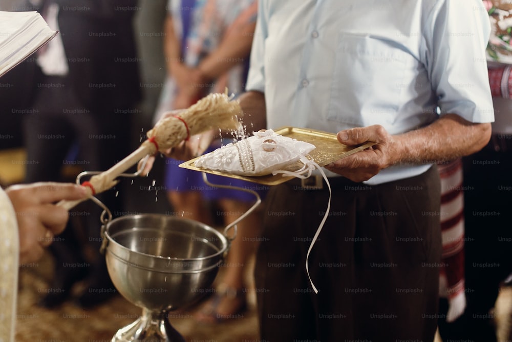Prêtre chrétien orthodoxe arrosant des alliances en or sur un oreiller blanc avec de l’eau bénite, rituel religieux sacré à l’église lors de la cérémonie de mariage