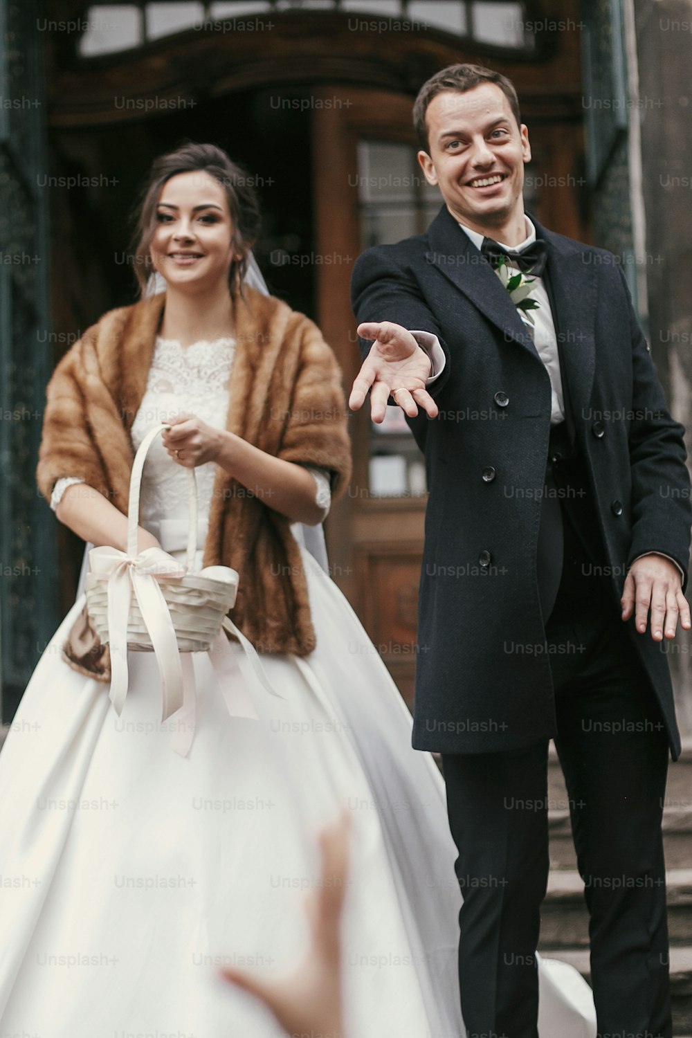 Hermosa novia con abrigo y elegante novio lanzando dulces cerca de la puerta de la iglesia después de la ceremonia de la boda.  feliz pareja de recién casados sonriendo y divirtiéndose. romántico 
 momento