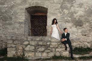 Handsome confident groom in stylish dark green suit with white bowtie sitting on stone steps near beautiful bride in white dress near castle entrance, romantic portrait