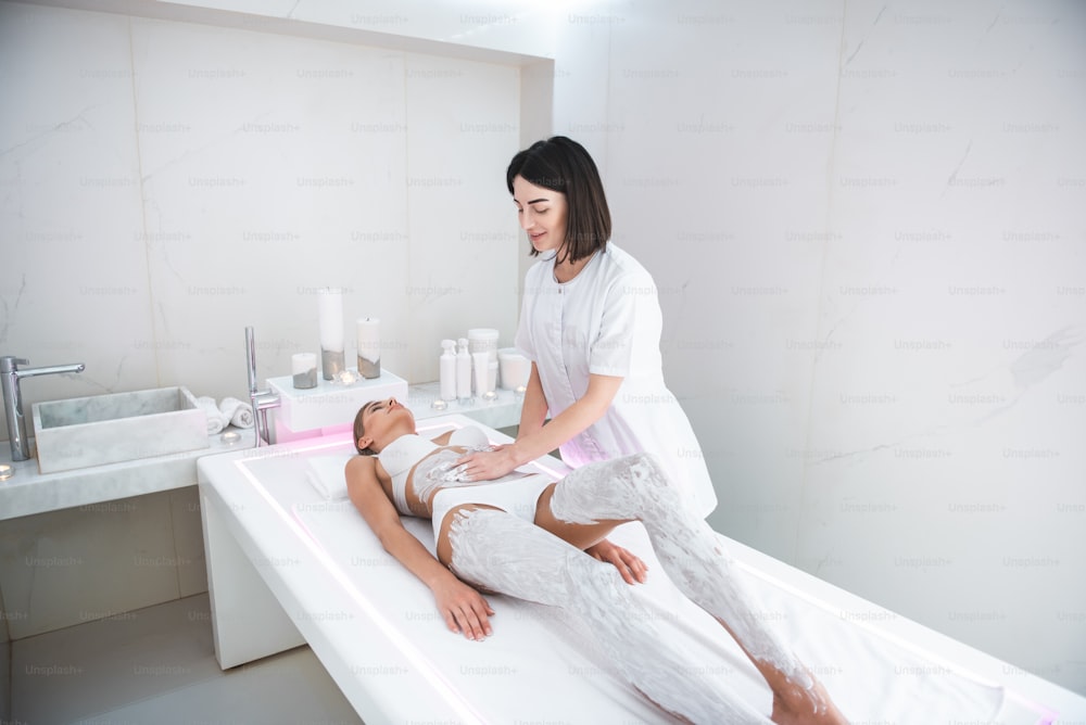 Complete relaxation. Smiling masseur doing spa procedure with cosmetic mask while lady lying on massage table