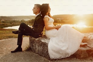 Romantic couple of newlyweds resting on stone bench at sunset field near lake landscape, happy bride leaning against smiling groom outdoors